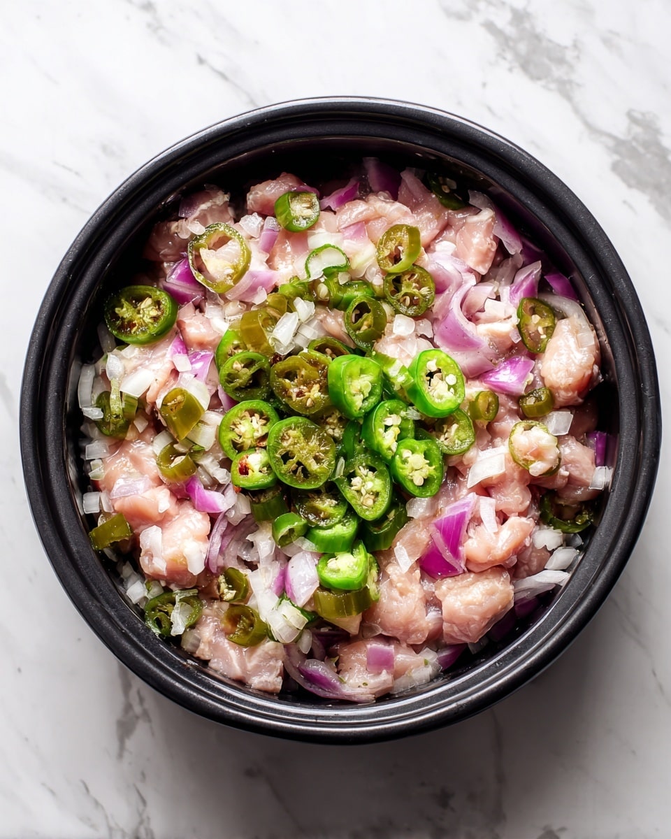 The image shows a black pot filled with diced light pink chicken pieces cooking on top of a layer of chopped onions that are translucent and soft. On top of the chicken and onions, there are small bright green sliced chili pieces scattered in the center. The pot edges curve gently, framing the cooking ingredients inside. The scene is set against a white marbled surface. photo taken with an iphone --ar 4:5 --v 7