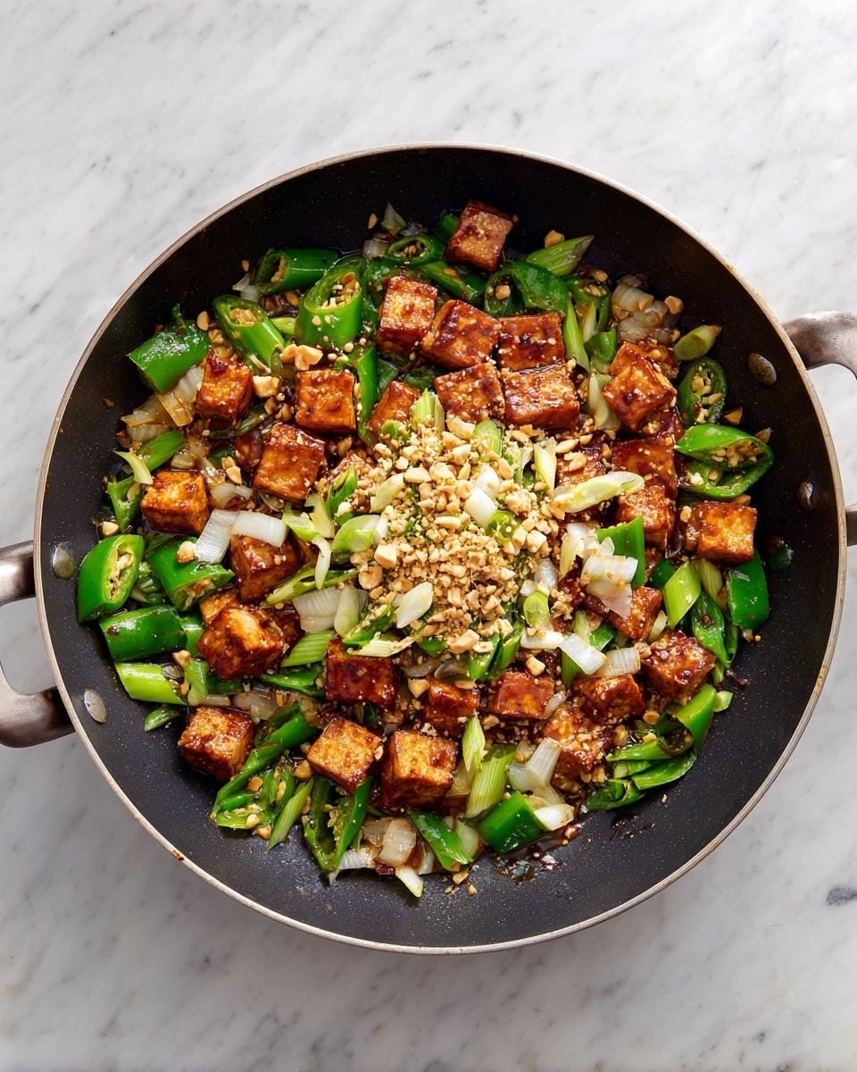 A dark pan holds a mix of crispy brown tofu pieces arranged in the center, surrounded by bright green chili slices and small bits of white onion scattered around the edges. Some crushed nuts are sprinkled on top of the tofu, adding texture and a light golden color. The tofu looks slightly browned and firm, contrasting with the fresh green and white elements. The pan is set on a white marbled surface. Photo taken with an iphone --ar 4:5 --v 7