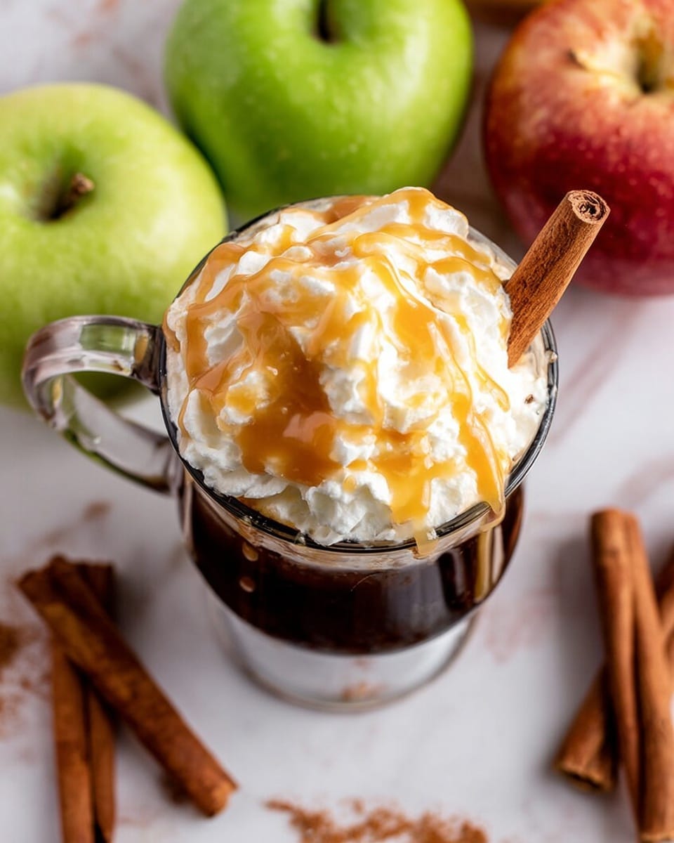 A clear glass cup filled with a dark liquid topped with a thick layer of white whipped cream, drizzled generously with golden caramel sauce. A cinnamon stick is placed on the right side of the whipped cream, partially inside the cup. Surrounding the cup are fresh apples with green and red tones, as well as cinnamon sticks scattered on a white marbled surface. Photo taken with an iphone --ar 4:5 --v 7