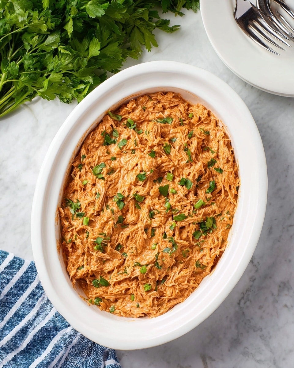 In the image, there is one layer of shredded chicken mixed with a creamy orange sauce, placed inside a white oval slow cooker dish. The chicken is covered evenly with the thick sauce that has a smooth, slightly chunky texture. Small green herbs are sprinkled on top for garnish, adding a pop of color. The dish sits on a white marbled surface with a bunch of fresh green cilantro on the upper left corner, a blue and white striped cloth near the bottom left, and two metal forks resting on a white round plate on the upper right. Photo taken with an iphone --ar 4:5 --v 7