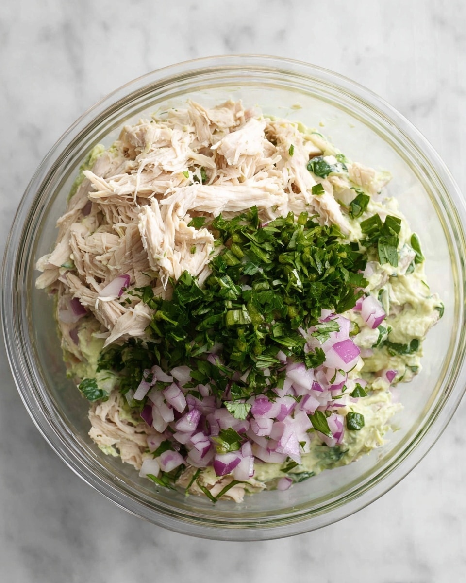A clear glass bowl shows a layered mix starting at the bottom with a pale green creamy layer, topped by a generous amount of shredded pale beige chicken. On top of the chicken, there is a mix of finely chopped light purple onions and bright green chopped herbs, adding fresh color contrast. The bowl is placed on a white marbled surface, giving a clean, fresh look. photo taken with an iphone --ar 4:5 --v 7