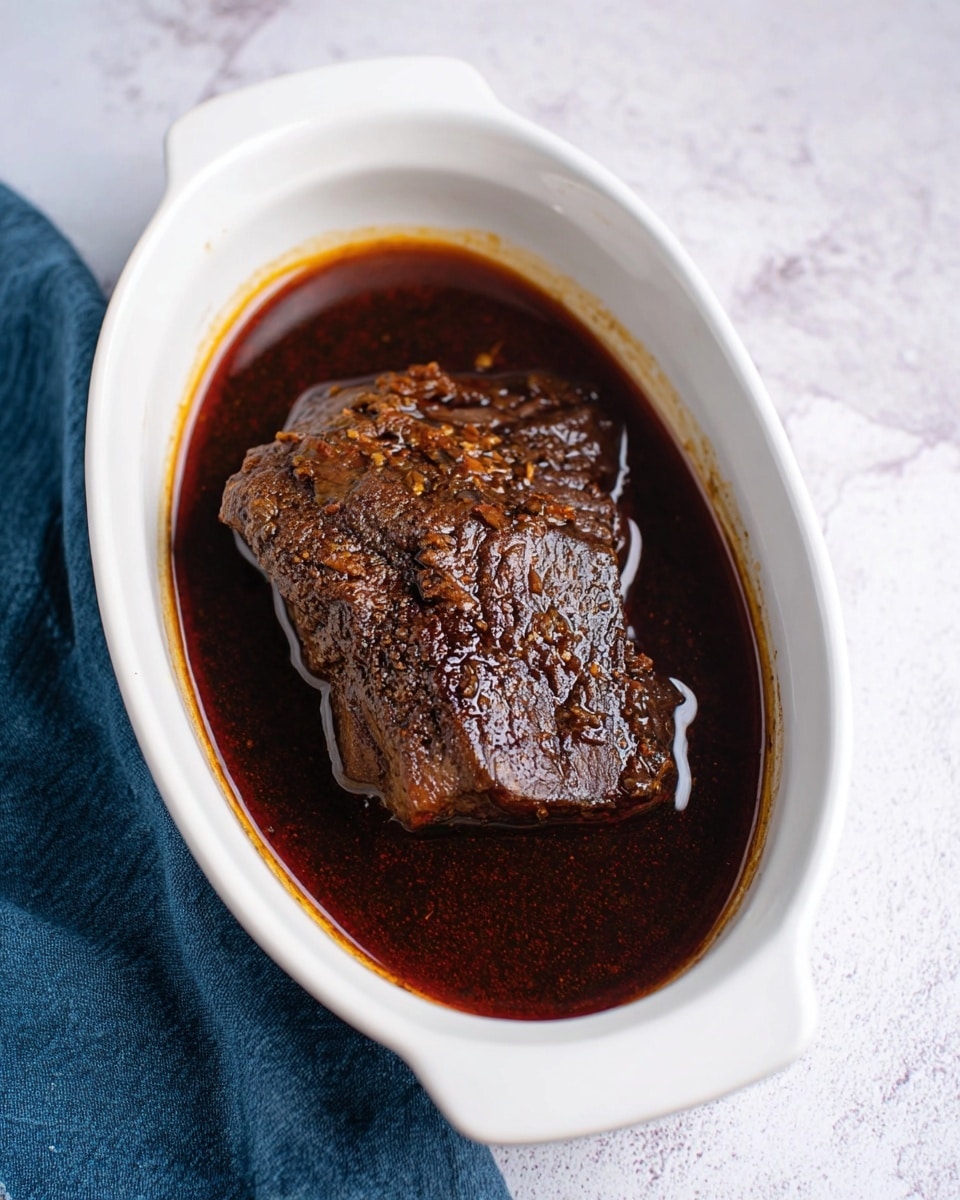 A white oval dish holds a single piece of dark brown cooked meat resting in a thick, shiny, dark sauce that fills the base of the dish. The meat has a textured surface with some burnt edges, and the sauce has a smooth, glossy look. The dish is placed on a white marbled textured surface with a blue cloth partially under the dish on the left side. Photo taken with an iphone --ar 4:5 --v 7