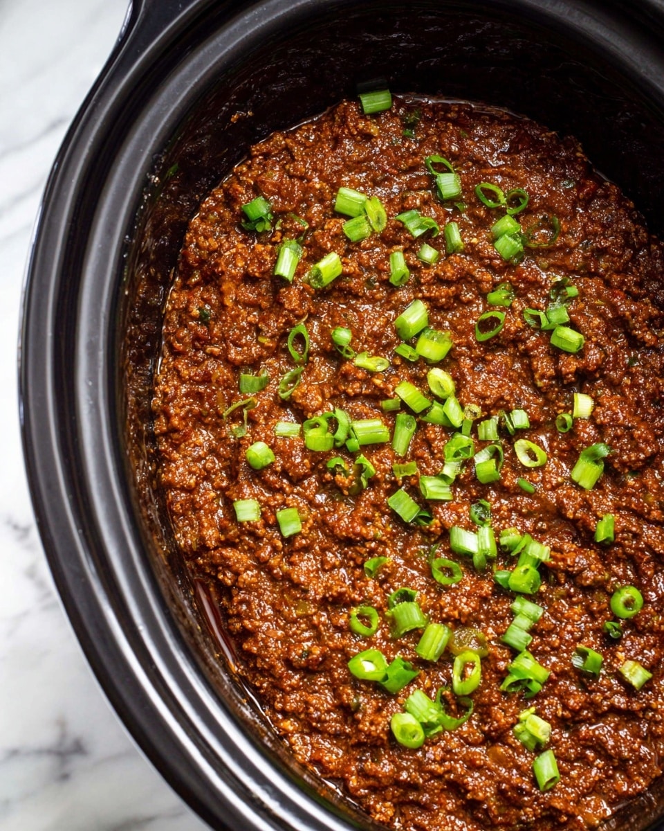 The image shows a close-up of a dark brown, thick meat sauce with a rich texture inside a black slow cooker. The sauce is evenly spread and topped with small pieces of bright green chopped scallions scattered across the surface. The slow cooker contrasts with the white marbled background visible around the edges. The sauce looks smooth but with small chunks, showing a glossy and moist appearance. Photo taken with an iphone --ar 4:5 --v 7