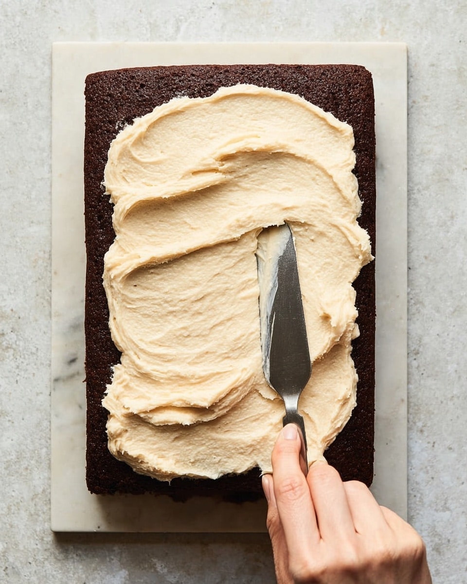 A dark brown rectangular cake sits flat on a white marbled surface. On top, there is one thick layer of creamy, light beige frosting being spread evenly over the cake with a metal spatula. A woman's hand holds the spatula, smoothing the soft, fluffy frosting from the center to the edges. The frosting's texture is creamy and slightly swirled as it is spread across the surface, contrasting with the smooth, baked texture of the cake below. Photo taken with an iphone --ar 4:5 --v 7
