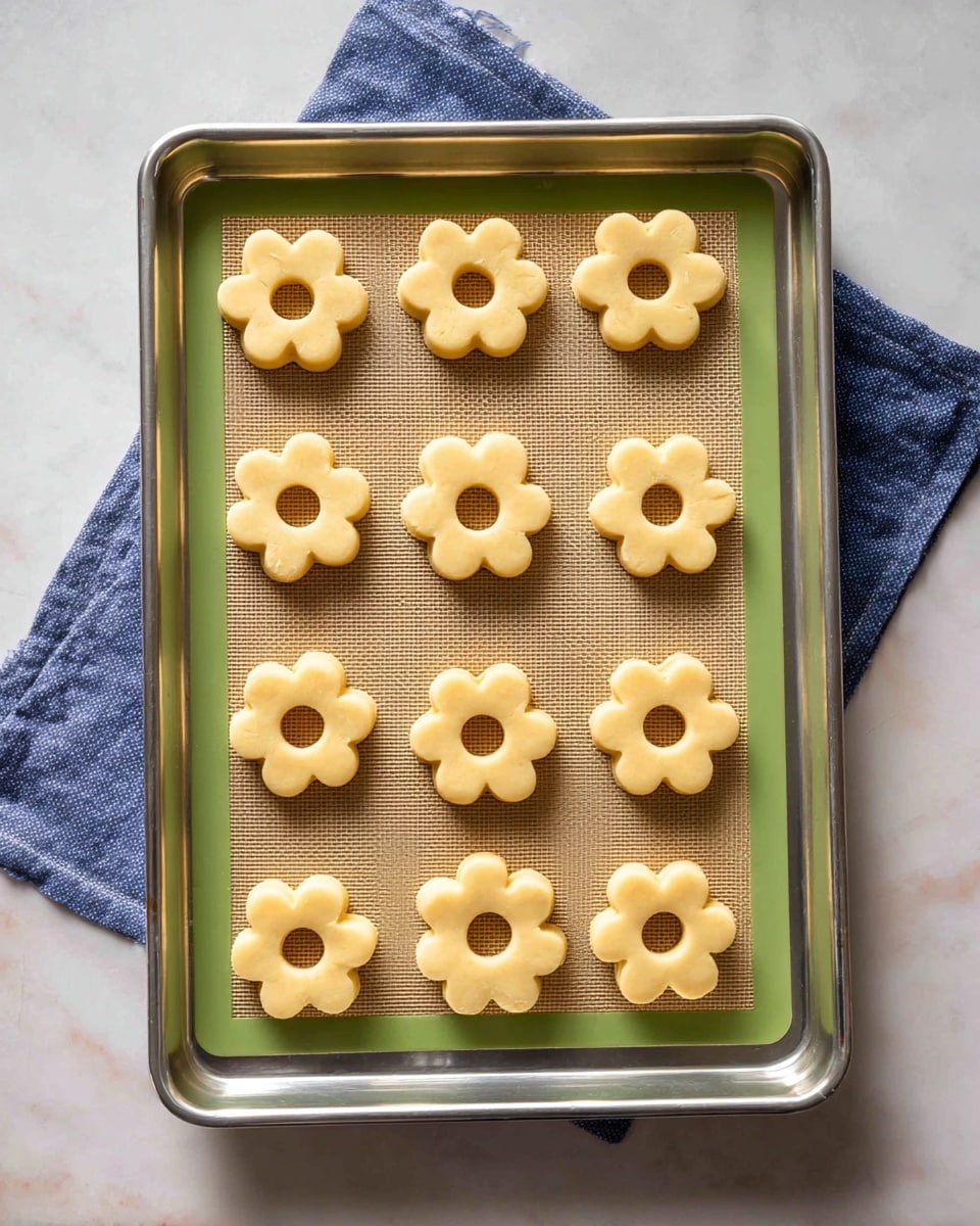 A silver baking tray holds a green-bordered silicone baking mat topped with twelve flower-shaped dough pieces arranged in a 4x3 grid. Each dough piece is a pale, creamy yellow color with a smooth texture and a round hole in the center. The tray rests on a white marbled surface, and a folded blue cloth is placed at the top edge. The overall look is clean and organized, with a soft, natural light illuminating the dough pieces evenly. photo taken with an iphone --ar 4:5 --v 7