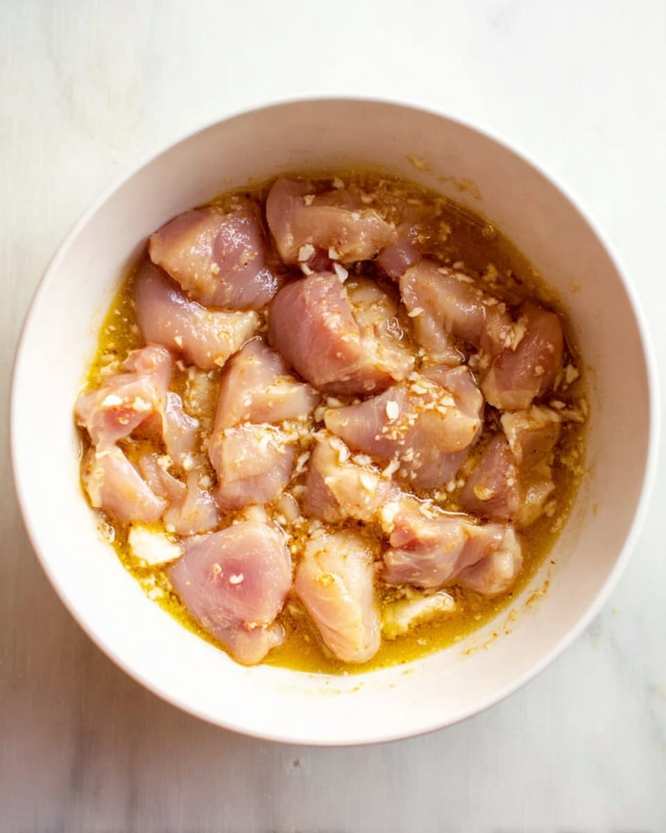 A white bowl filled with small, uneven pieces of raw light pink chicken mixed with a slightly thick, golden yellow marinade, some pieces showing a glossy, moist texture with tiny white bits scattered around, the marinade has a few bubbles and streaks of oil, all sitting on a white marbled surface photo taken with an iphone --ar 4:5 --v 7