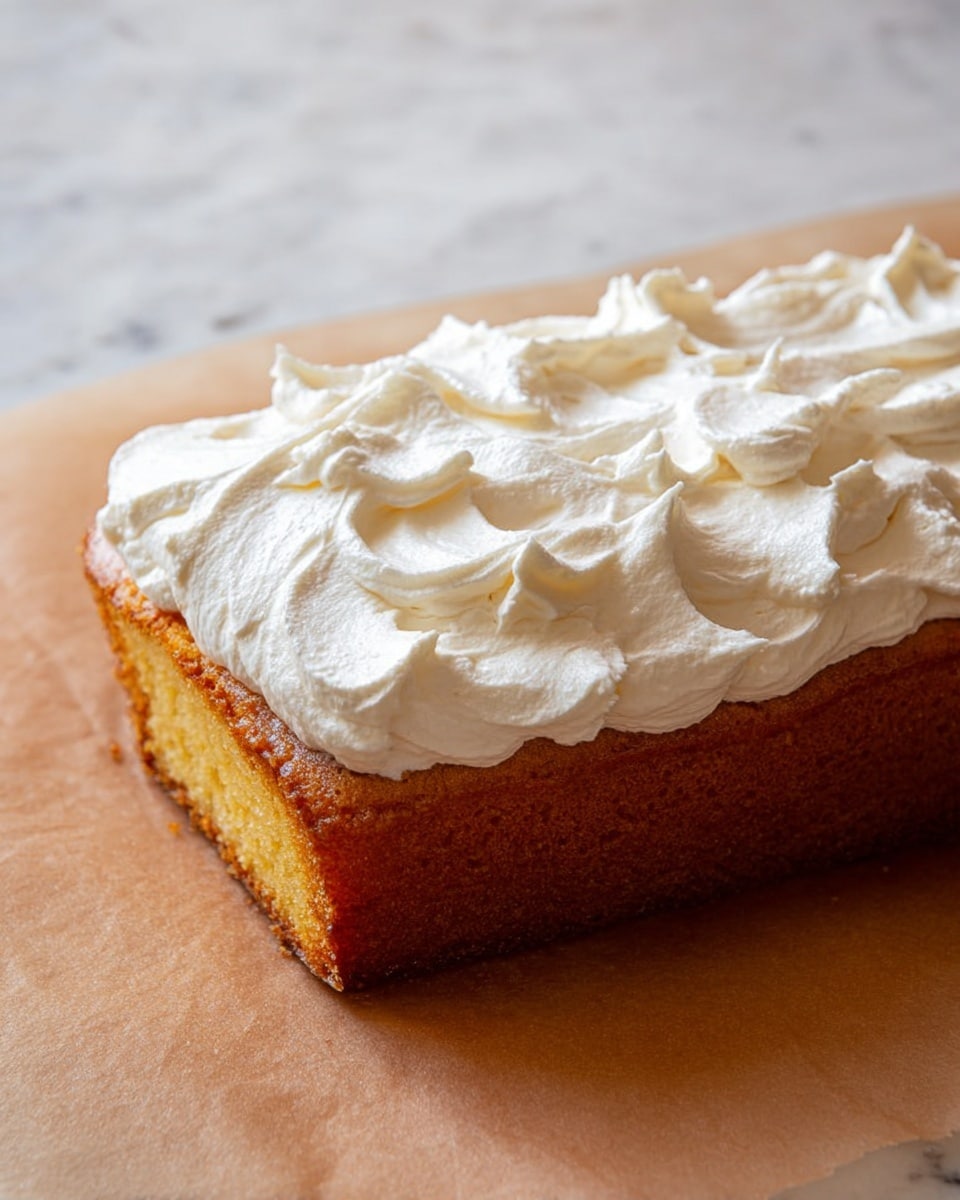 The image shows a single rectangular piece of yellow cake with a thick layer of white whipped cream frosting on top. The frosting is spread unevenly with soft peaks and swirls, creating a fluffy and textured look. The cake has a golden-brown crust on the side and sits on a tan parchment paper placed on a white marbled surface. Photo taken with an iphone --ar 4:5 --v 7
