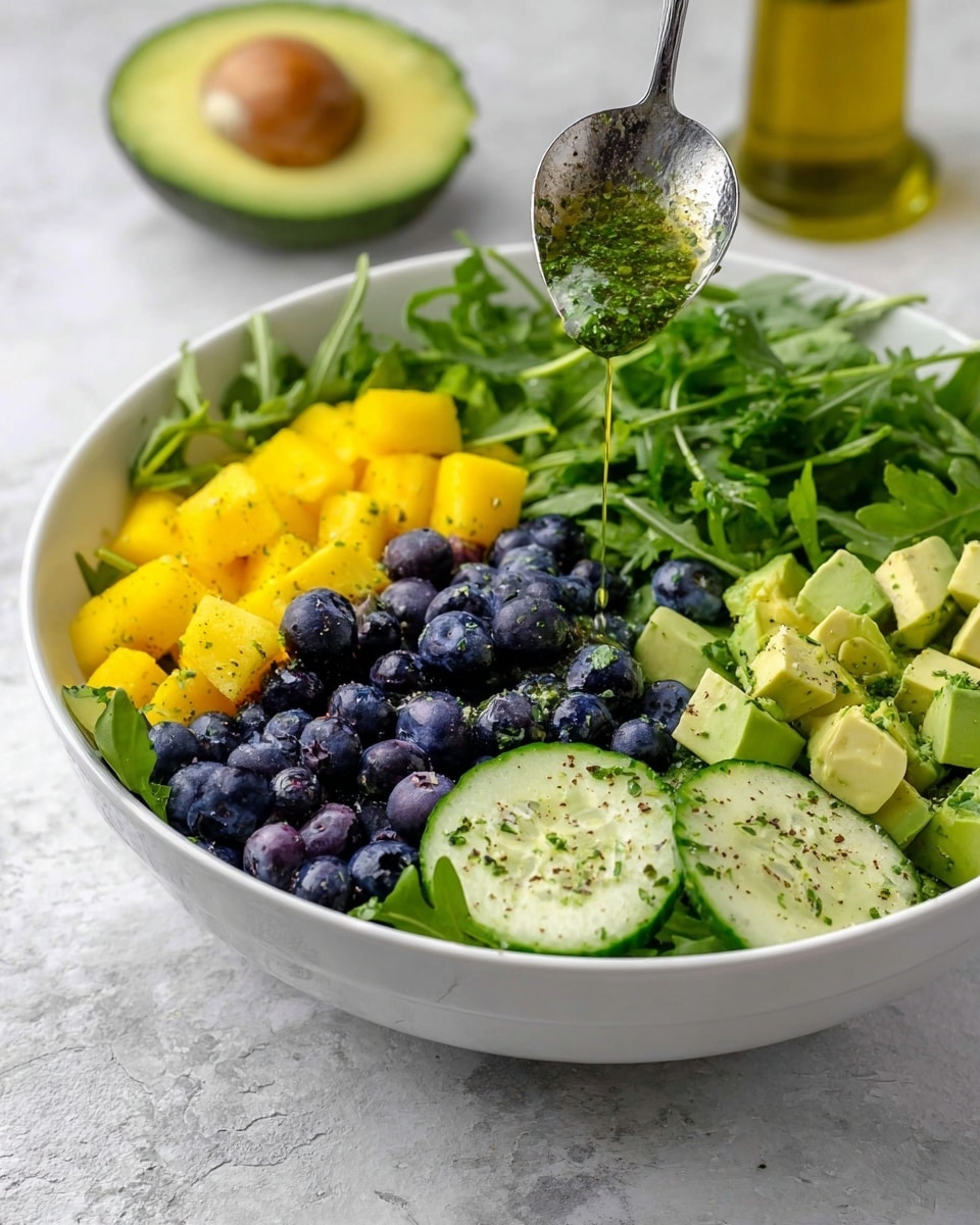 A white bowl contains a colorful layered salad on a white marbled surface. The bottom layer is fresh green arugula and parsley leaves covering half the bowl. On the left side, there is a bright yellow layer of cubed mango. In the center, there are fresh, round blueberries with a shiny texture. Surrounding the blueberries on the right are light green, cubed avocado pieces with some herbs sprinkled on top. Below the blueberries, round slices of cucumber with visible seeds are placed, lightly sprinkled with black pepper. A silver spoon is held above the bowl, pouring a green herb dressing onto the salad. In the background, a halved avocado and a glass bottle of olive oil are visible. Photo taken with an iphone --ar 4:5 --v 7