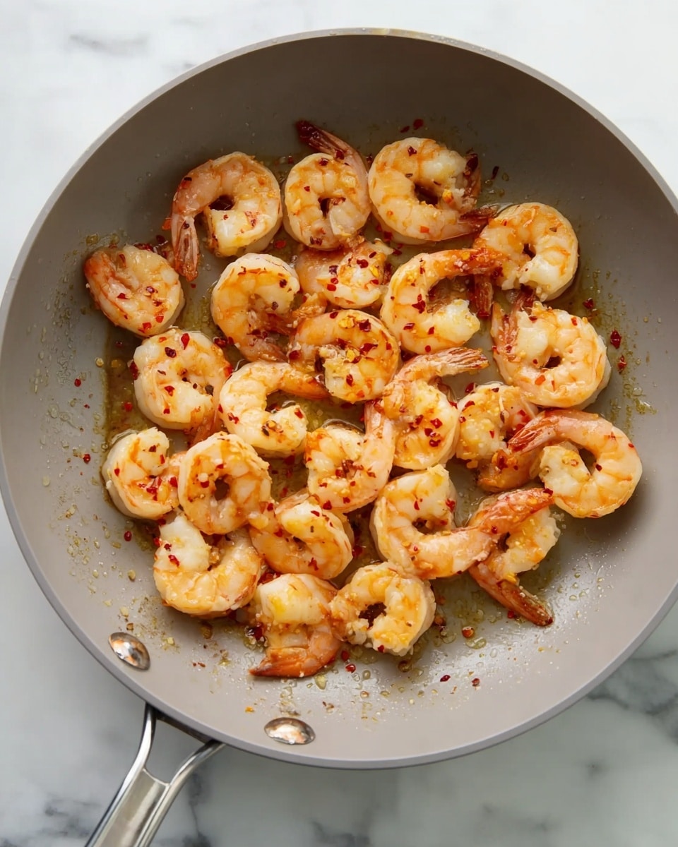 A gray frying pan filled with a single layer of cooked shrimp with a light orange color and some red chili flakes scattered on top. The shrimp are curled and evenly spread inside the pan. The pan sits on a white marbled surface, showing some small spots of oil. The pan handle is visible on the left side with a metal loop on the right side. photo taken with an iphone --ar 4:5 --v 7