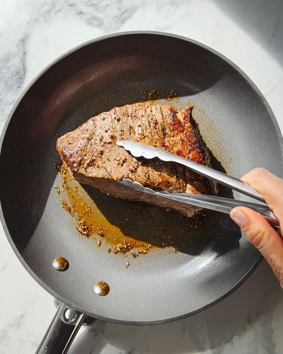 A large piece of browned cooked meat is being held with silver tongs by a woman's hand inside a gray non-stick pan, with a small bit of sauce or seasoning underneath the meat. The surface under the pan is white with a marbled texture. The lighting is bright and natural, highlighting the color and texture of the meat. photo taken with an iphone --ar 4:5 --v 7