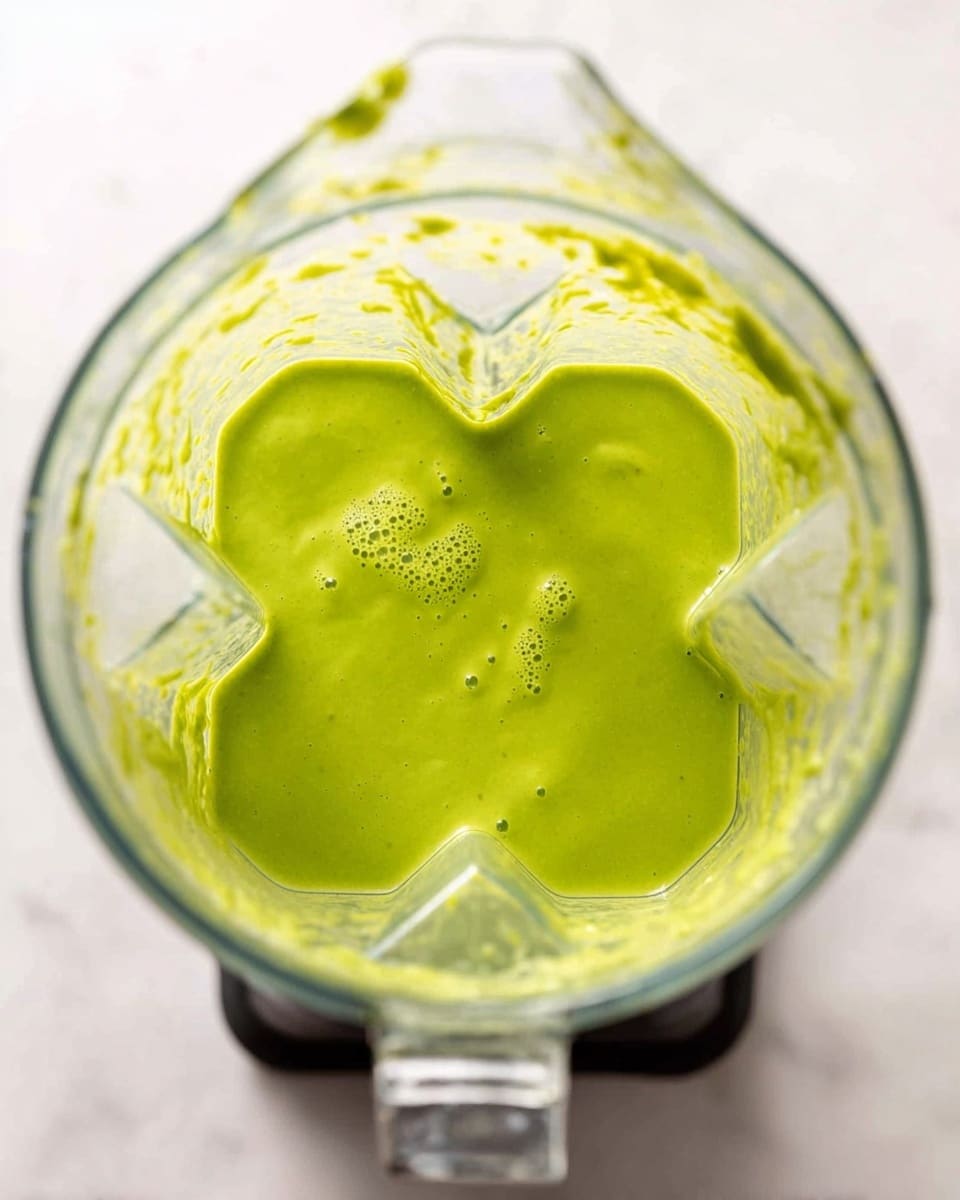 A clear blender pitcher filled with a smooth, creamy green sauce or dressing, with a bright and fresh color and a few small bubbles on the surface showing its light texture. The inside of the blender has some sauce splattered around its clear sides, and the blades are visible beneath the sauce at the bottom. The blender is set on a white marbled surface which contrasts softly with the vibrant green sauce. photo taken with an iphone --ar 4:5 --v 7