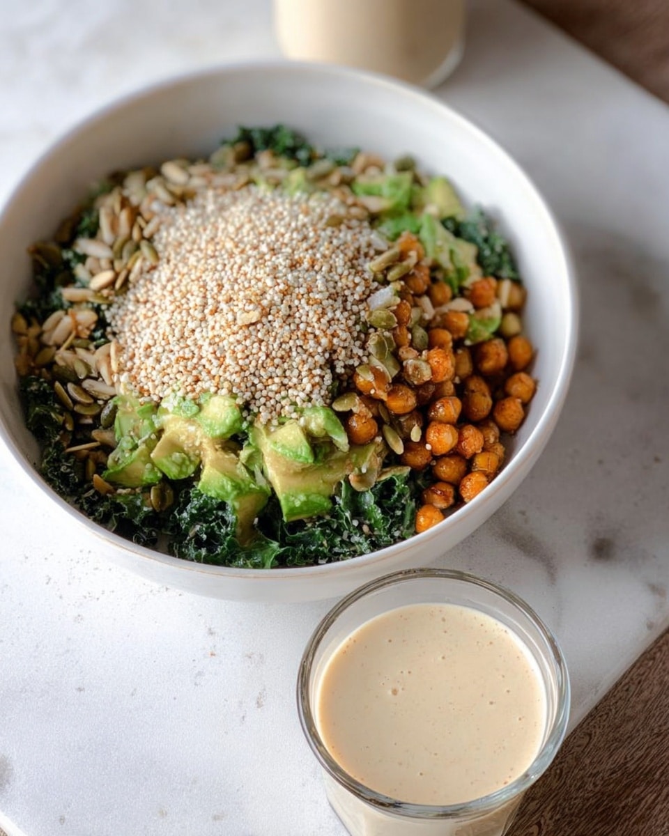 A white bowl sits on a white marbled surface containing a layered salad. The bottom layer shows dark green leafy kale, topped with light brown roasted chickpeas. Next to this is a layer of bright green avocado chunks on one side. The top layer is covered with a thick pile of beige sesame seeds and some pumpkin seeds. In front of the bowl, there is a small clear glass cup filled with a light beige creamy sauce. photo taken with an iphone --ar 4:5 --v 7