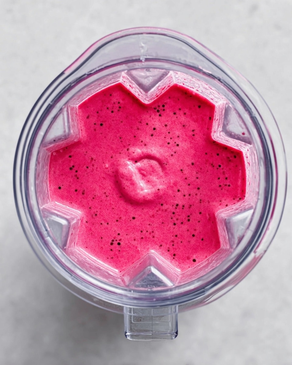 A top view of a blender container filled with a smooth, bright pink liquid with tiny black specks scattered throughout. The inside of the clear blender container shows sharp edges and clear plastic details, contrasting with the vibrant pink color of the mixture. The blender sits on a white marbled surface, highlighting the intense color of the blend inside. photo taken with an iphone --ar 4:5 --v 7