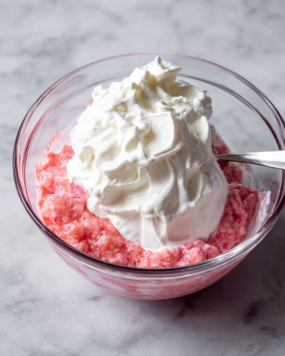 The image shows a clear glass bowl filled with a bright pink, chunky mixture at the bottom layer. On top of this is a thick, smooth, white cream layer that looks fluffy. The cream is piled high in soft peaks, covering part of the pink mixture beneath. There is a silver spoon sticking into the cream from the right side of the bowl. The bowl sits on a white marbled surface. photo taken with an iphone --ar 4:5 --v 7