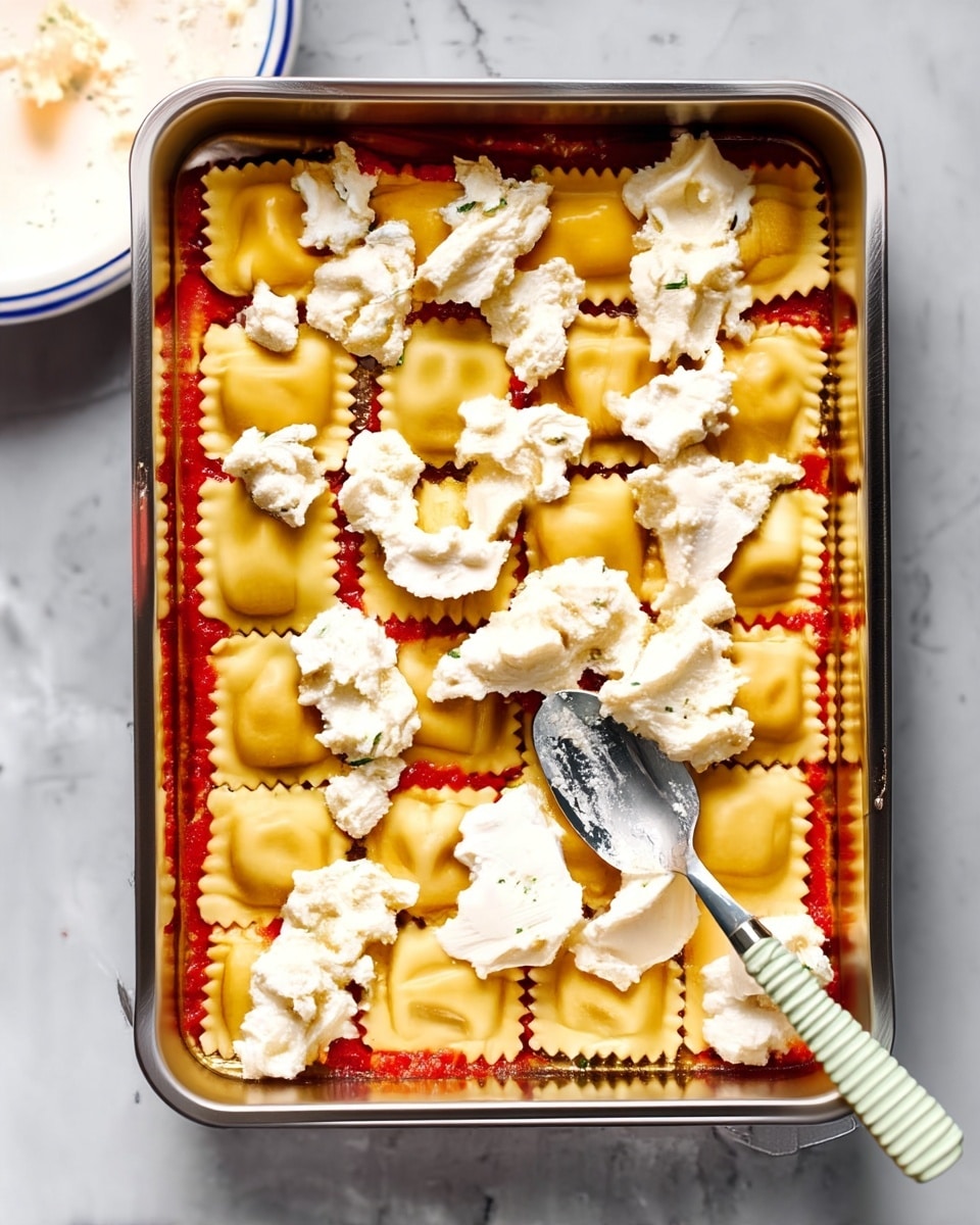 A silver rectangular baking dish holds a layer of red tomato sauce evenly spread at the bottom. On top, there is a single layer of square yellow ravioli arranged neatly, filling the dish edge to edge. Dollops of white, creamy ricotta cheese are scattered unevenly across the ravioli, some larger and some smaller. A spoon with a light green and white striped handle rests on the right side of the dish, with some cheese on it. The dish sits on a white marbled surface, and a white plate with blue stripes around the edge is partially visible in the top left corner, containing more white cheese. Photo taken with an iphone --ar 4:5 --v 7