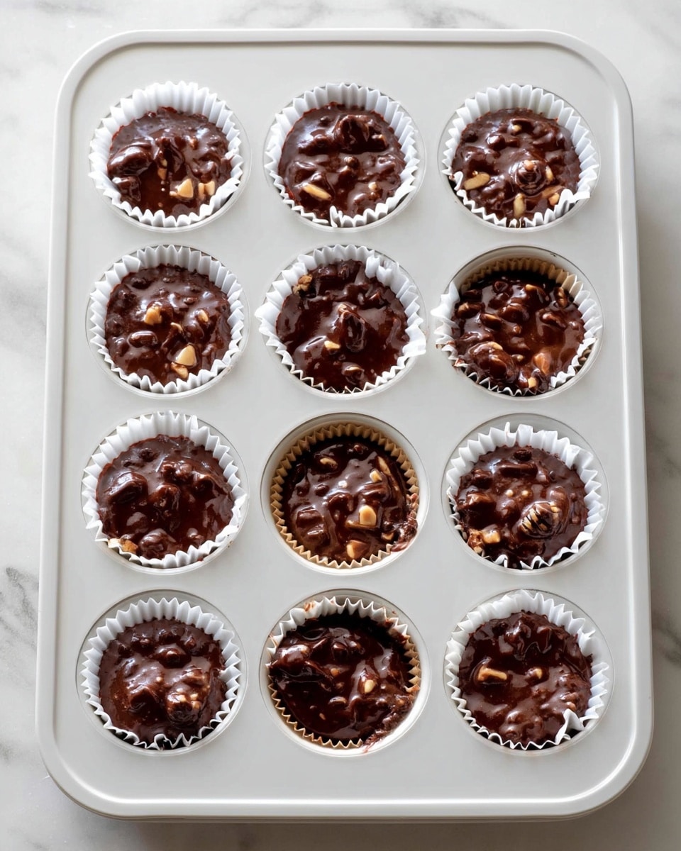 A white muffin tray holds 12 cups, each filled with dark brown chocolate batter mixed with small chunks of nuts or chocolate. The batter has a thick and slightly lumpy texture, filling each white paper liner almost to the top. The tray is placed on a white marbled surface, showing some soft natural light and shadows. photo taken with an iphone --ar 4:5 --v 7
